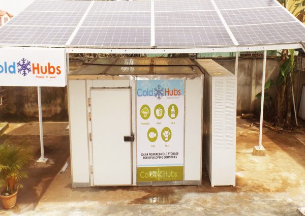 A solar-powered cold storage unit with a metal roof covered in solar panels, exemplifying renewable energy. The unit, labeled "ColdHubs," displays icons for freshness, saving money, reducing waste, and constant temperature. It is situated on a concrete surface outdoors.